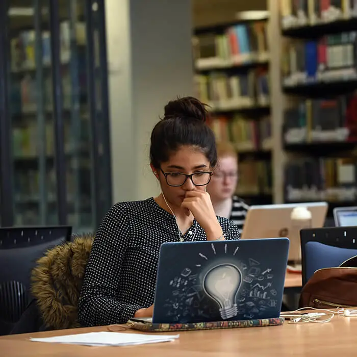 female student with headphones watching a video on her laptop in the university library with other student in the background
